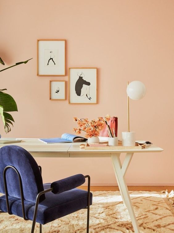 a peachy pink home office with a white desk, a navy chair, a small gallery wall and some blooms and greenery