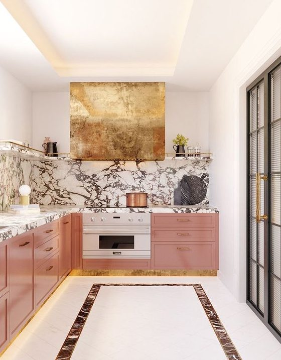 a pink kitchen with a white stone backsplash and countertops plus a shiny gold hood for a super glam accent