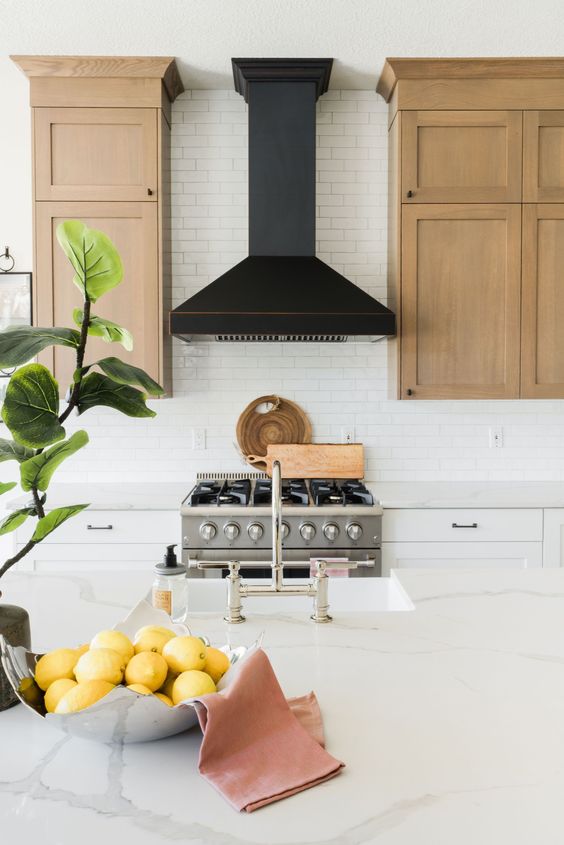A pretty airy kitchen with light stained and white cabinets, a white tile backsplash, a black hood and a large kitchen island
