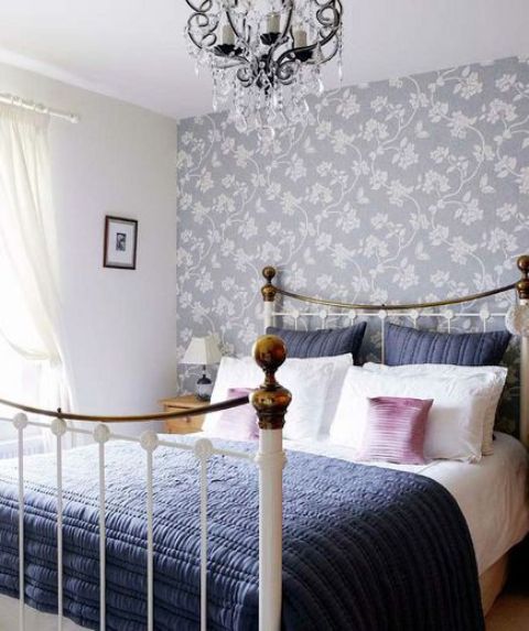 a pretty glam bedroom with a grey floral accent wall, a metal bed with navy and white bedding, nightstands, a crystal chandelier