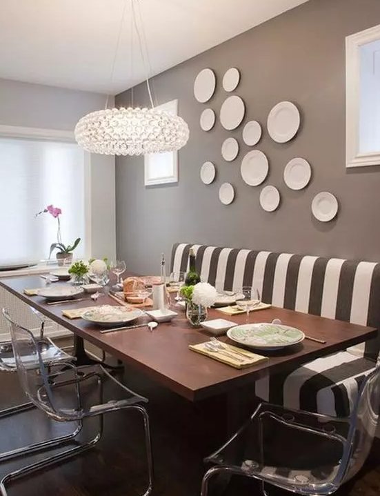 a quirky taupe dining room with a striped sofa, a wooden table, acrylic chairs, a chic chandelier and a gallery wall of plates