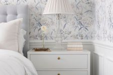a refined and subtle bedroom with blue floral wallpaper, a pale blue bed with neutral bedding, a white nightstand, a white lamp