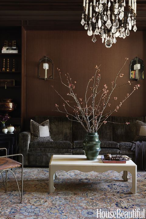 A refined living room with brown walls and built in shelves, a grey sofa, a crystal chandelier, a coffee table and some chairs
