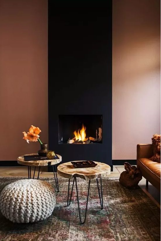 A refined modern living room with a rust colored wall and a black fireplace, a leather sofa, wood slice tables and a knit ottoman