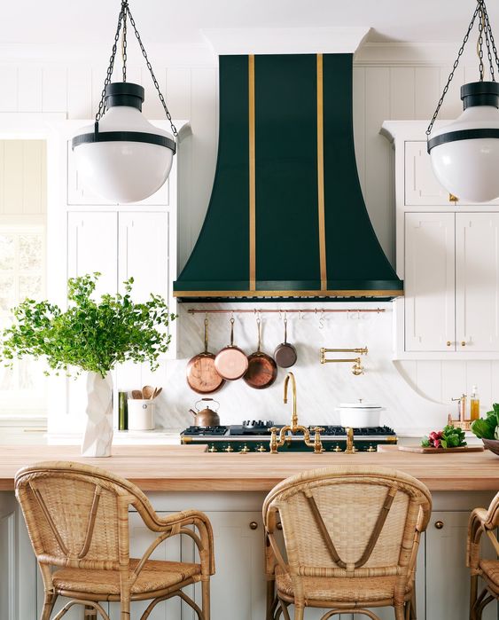 a refned white vintage kitchen with a dark green and gold hood over the cooker and a kitchen island with rattan chairs