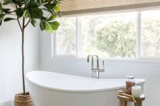 a serene neutral bathroom with white walls and a tan herrinbgone tile floor, an oval tub and a wooden stool, a basket and a potted tree