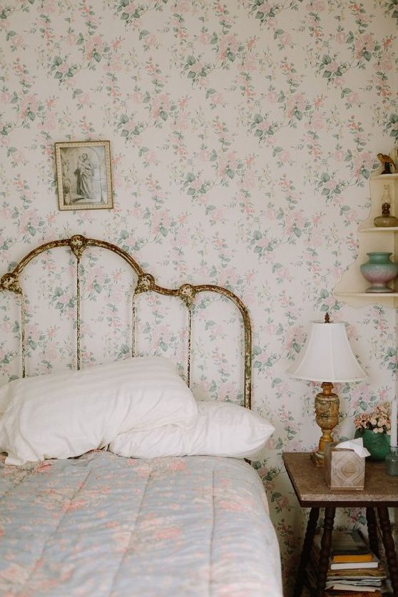 a shabby chic bedroom with delicate floral wallpaper, a shabby chic bed, floral bedding, a stool as a nightstand and a vintage lamp