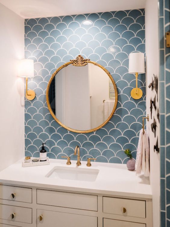 a sink space with a blue scallop tile accent wall, a neutral vanity, a round mirror, brass fixtures and cool wall lamps