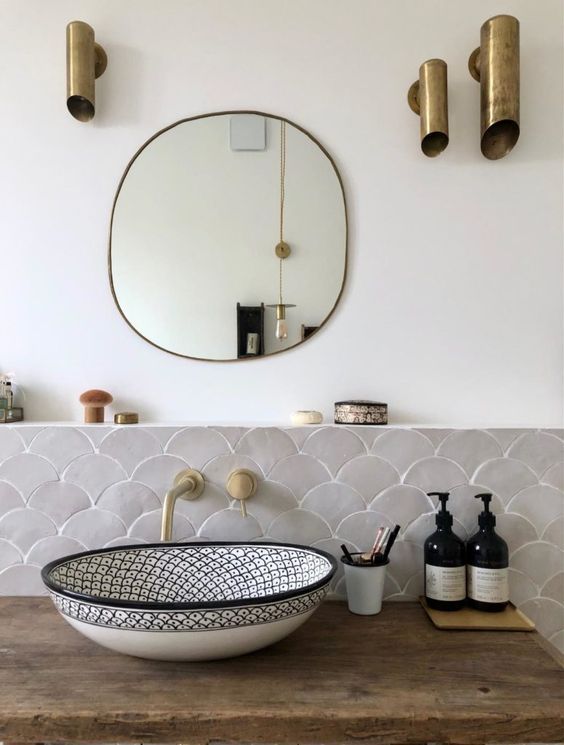 a small and elegant sink space with grey scallop tiles, a printed sink, a mirror and catchy brass tube lamps