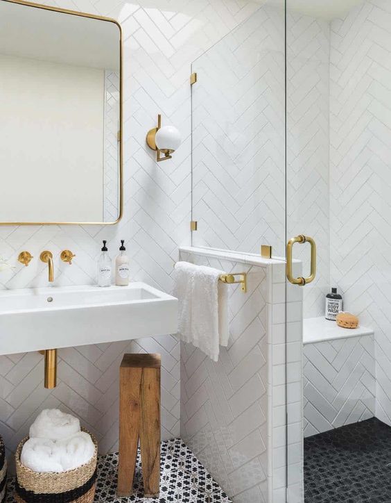 A small bathroom clad with white herringbone tiles, a shower space with a bench, a wall mounted sink and gold fixtures