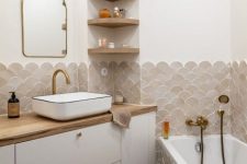 a small neutral bathroom done with greige and beige fishscale tiles, a bathtub clad with the same tiles, a white vanity with butcherblock countertops