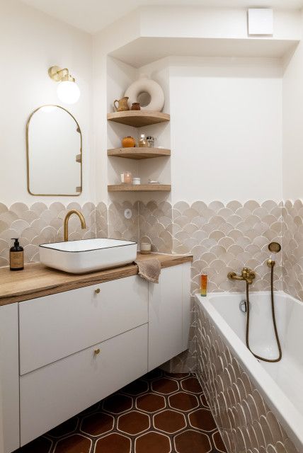 a small neutral bathroom done with greige and beige fishscale tiles, a bathtub clad with the same tiles, a white vanity with butcherblock countertops