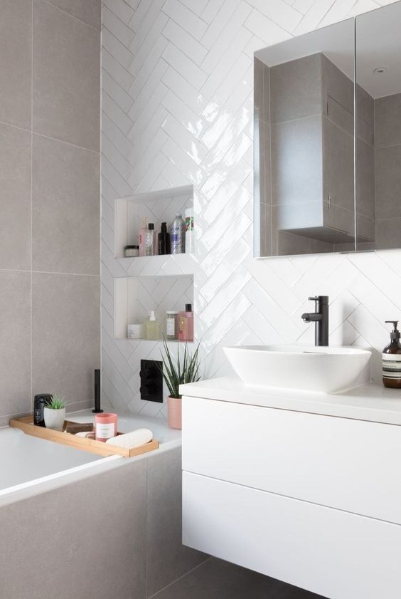 a small neutral bathroom with white herringbone tiles, grey stone ones, a white vanity, a mirror cabinet and niche shelves