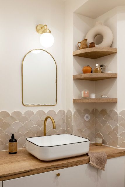 a small neutral space done with tan and beige fishscale tiles, a sink, open shelves with lovely decor and a mirror in a gilded frame