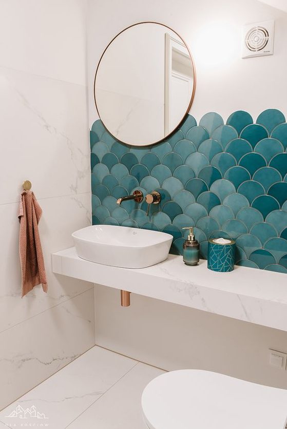 A small sea inspired bathroom clad with white marble tiles and blue and turquoise fish scale ones, a vanity with a sink and a round mirror