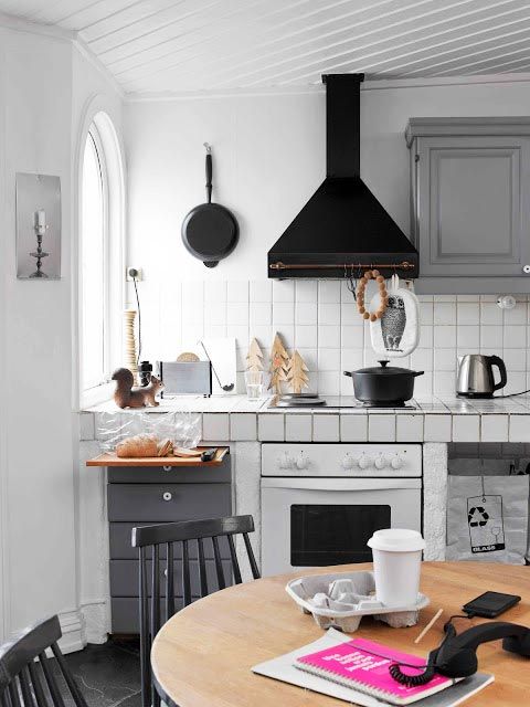 a small white Scandinavian kitchen with white tiles all over, some grey cabinets, a black hood and some black chairs