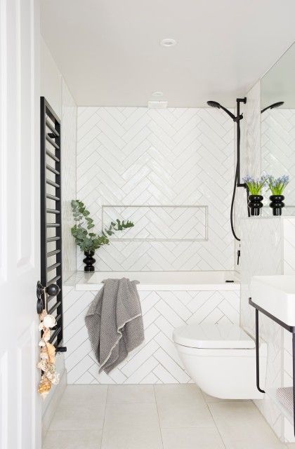 A small white bathroom done with herringbone tiles, a free standing vanity, black fitures and greenery