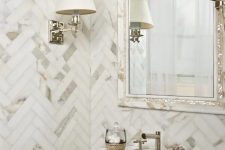a small yet luxurious marble powder room with chevron clad walls and a chic mirror plus wall lamps