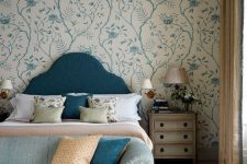 a sophisticated bedroom with a blue floral accent wall, a navy bed with neutral bedding, a shabby chic nightstand, some lamps and a ligth blue sofa