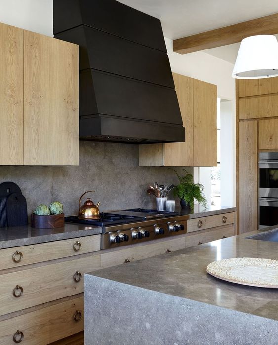 a stained kitchen with metal pulls, grey stone countertops and a backsplash, a large black hood is laconic and chic