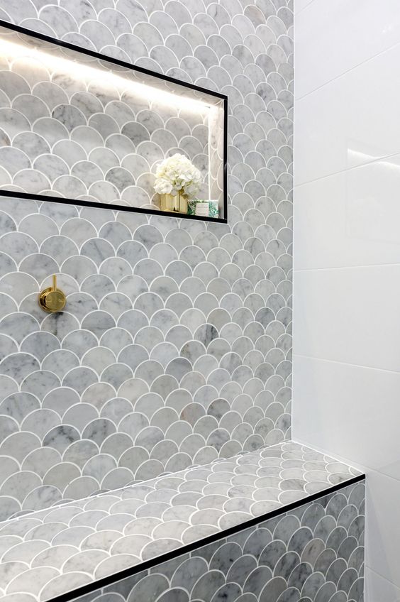 a stylish grey and white bathroom with large scale and grey scallop tiles, a niche shelf with lights and gold fixtures