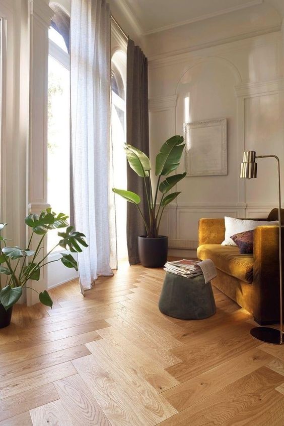 A stylish off white living room with a herringbone floor, a mustard chair, a green pouf, potted plants and a gold floor lamp