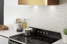 a stylish white kitchen with white terrazzo countertops and a backsplash, a shiny gold hood over the black cooker