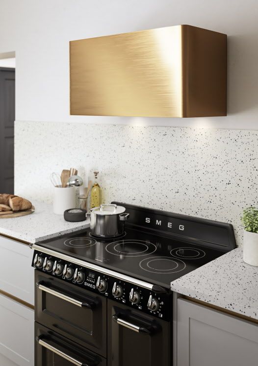 a stylish white kitchen with white terrazzo countertops and a backsplash, a shiny gold hood over the black cooker