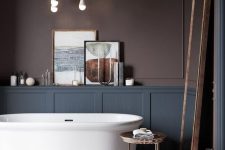 a super refined moody bathroom with chocolate brown walls, grey paneling, a chic tub on a marble platform and a floor mirror