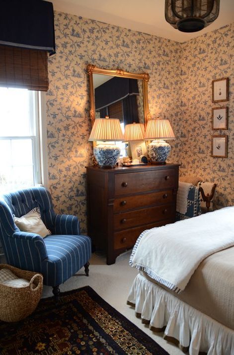 A vintage English bedroom with a bed and neutral bedding, a dark stained dresser, a blue chair, lovely blue wallpaper