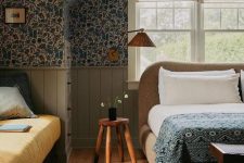 a vintage attic bedroom with dark floral wallpaper, a boucle upholstered bed with printed bedding, a rug and a sofa at the window