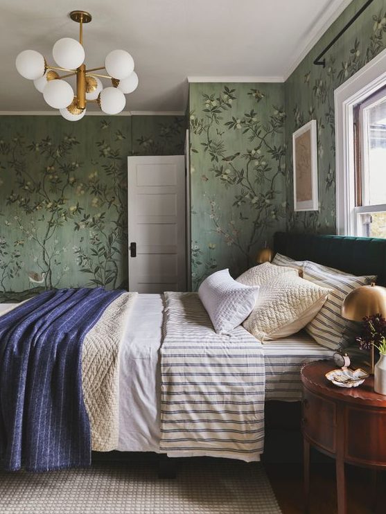 A vintage inspired bedroom with green floral wallpaper, a bed with printed bedding, a rich stained nightstand with a gilded lamp