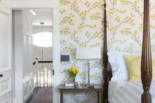 a vintage-inspired bedroom with yellow floral walls, a heavy bed with pillars, a chair and nightstands plus blooms