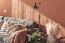a welcoming boho living room with a terracotta accent wall, a grey sofa with pillows, a carved boho side table, pampas grass and a floor lamp