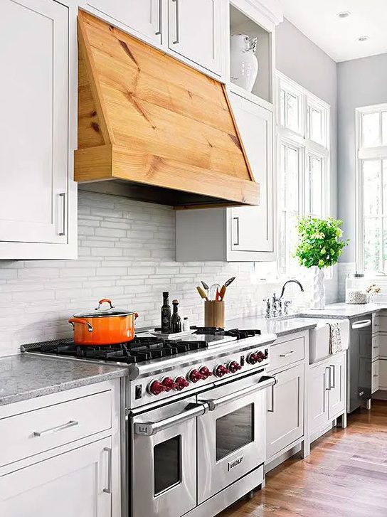 a white farmhouse kitchen with a white tie backsplash and stone countertops, a stained hood over the cooker that brings warmth