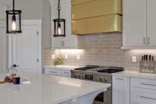a white farmhouse kitchen with shaker cabinets, a grey tile backsplash, a shiny gold hood and a small black pendant lamps