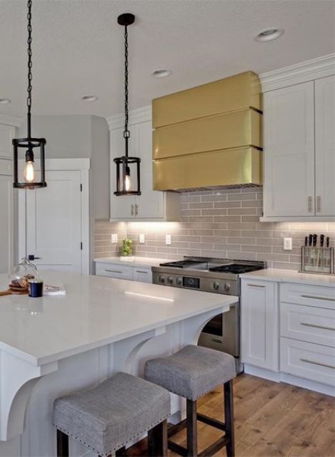 a white farmhouse kitchen with shaker cabinets, a grey tile backsplash, a shiny gold hood and a small black pendant lamps