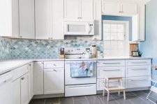 a white kitchen with a blue accent wall, shaker cabinets, a blue, aqua and white scallop tile backsplash and a gilded lamp