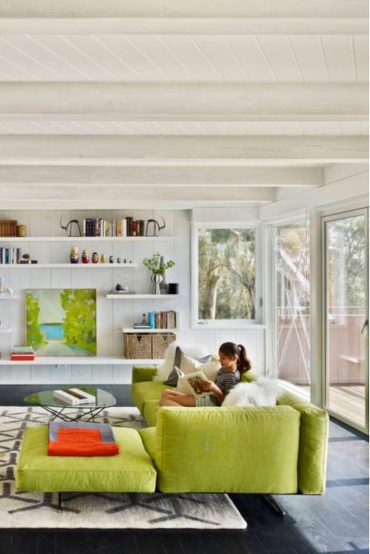 a white living room with beams on the ceiling, a chartreuse sectional, a glass coffee table and a printed rug