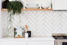 a white minimalist kitchen with a herringbone tile backsplash, open shelves and a sleek white kitchen island, some greenery