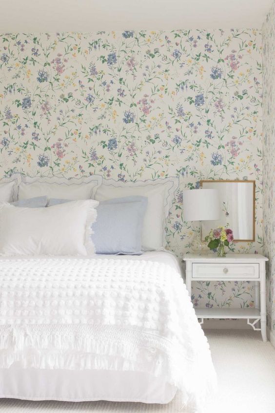 an English cottage bedroom with lovely pastel floral wallpaper, a bed with white and pastel blue bedding, a white nightstand and a mirror
