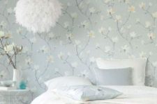 an airy bedroom with a blue floral wallpaper wall, a bed with neutral bedding, a metal nightstand, a basket and a fluffy pendant lamp