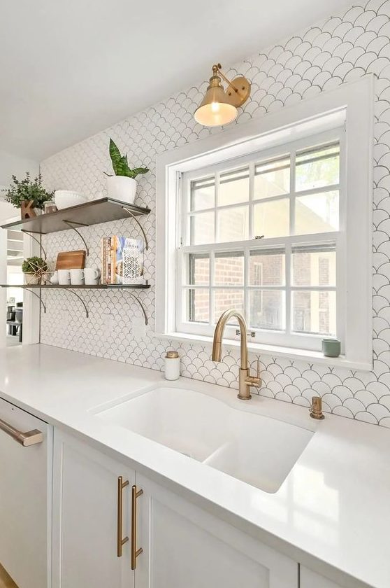 an airy white kitchen with shaker cabinets, a white fishscale tile backsplash, open shelves and brass fixtures and lamps