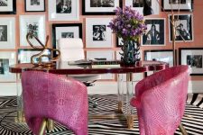 an artistic home office with a peachy pink accent wall fully covered with a black and white gallery wall, a unique desk and hot pink leather chairs on brass legs