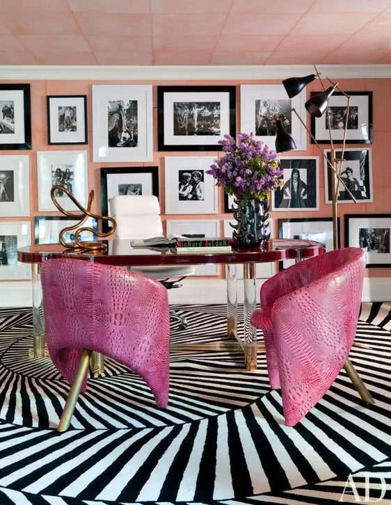 an artistic home office with a peachy pink accent wall fully covered with a black and white gallery wall, a unique desk and hot pink leather chairs on brass legs