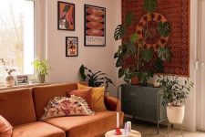 an earthy tone boho living room with some artwork, a rust-colored sofa, poufs and stools and potted greenery