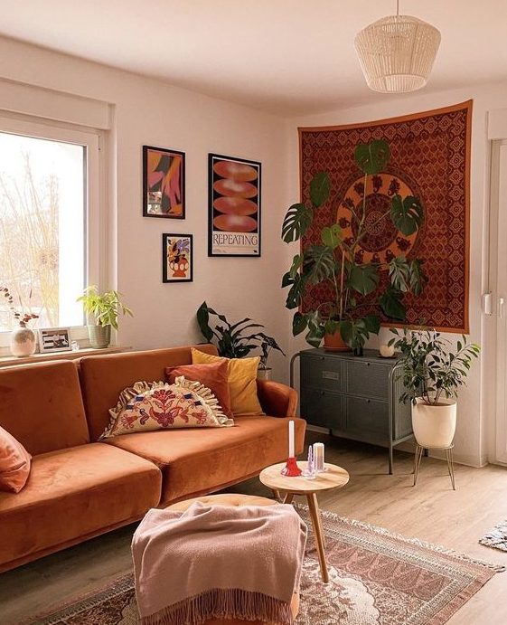 An earthy tone boho living room with some artwork, a rust colored sofa, poufs and stools and potted greenery