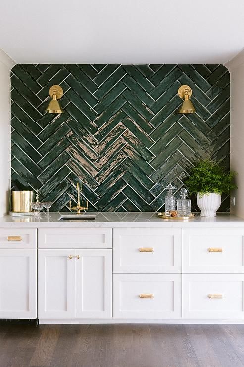 an elegant home bar with a green herringbone tile backsplash, white shaker cabinets, gold touches is very chic and elegant
