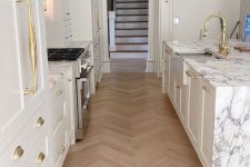 an exquisite white kitchen with shaker style cabinets, a large kitchen island with a white stone countertop and a light-stained herringbone floor