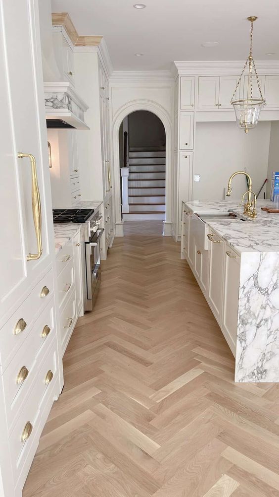 An exquisite white kitchen with shaker style cabinets, a large kitchen island with a white stone countertop and a light stained herringbone floor